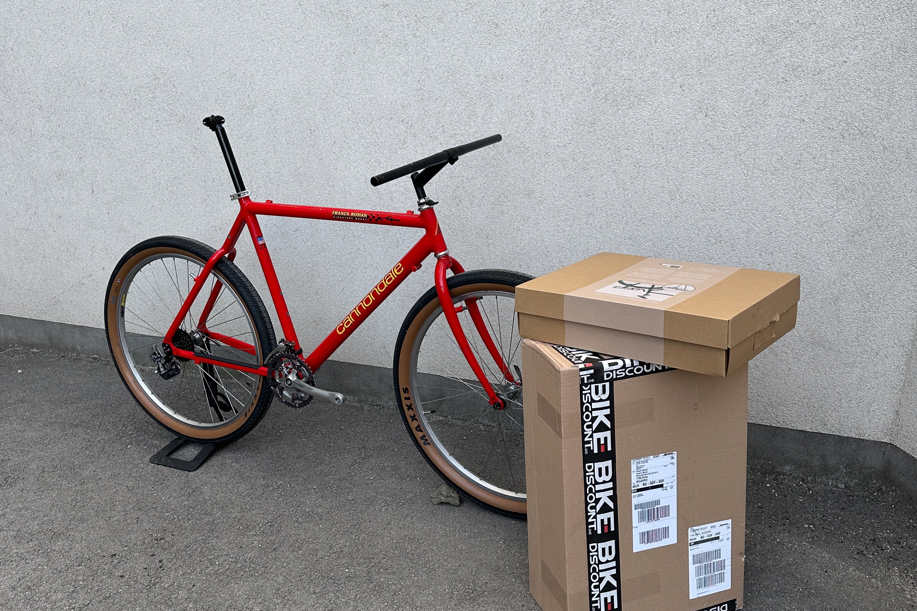 Red bicycle next to a cardboard box on a gray floor with a white wall background
